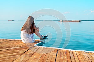 Lonely young girl in white dress sitting on the pier hugging her knees looking into the horizon, loneliness, parting, waiting, aut