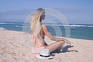 Lonely young woman sitting on the tropical beach by the sea with hat and in the swimsuit. Bali island vacation