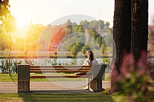 Lonely young woman sitting on a bench in the park near the lake on sunset