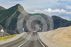 A long asphalt road with a white and yellow dividing strip against the backdrop of high mountains with rocks and stones on a clear