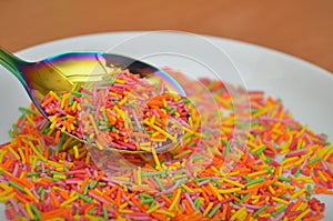 Long baking dots, sprinkle in a white saucer in rainbow spoon on a wooden table, decoration for bakery, closeup