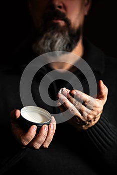 Long bearded man holding beard wax, balm or lotion, black background