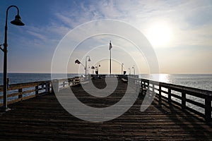 A long brown wooden pier with American flags flying on curved light posts with people walking and fishing on the pier