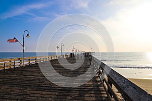 A long brown wooden pier with American flags flying on curved light posts with people walking and fishing on the pier