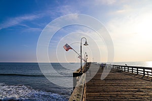 A long brown wooden pier with American flags flying on curved light posts with people walking and fishing on the pier