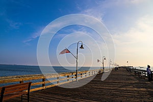 A long brown wooden pier with American flags flying on curved light posts with people walking and fishing on the pier