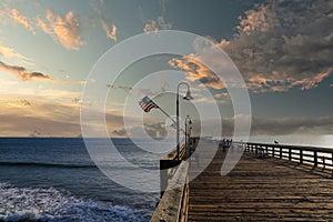 A long brown wooden pier with American flags flying on curved light posts with people walking and fishing on the pier