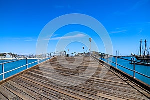 A long brown wooden pier with a gray metal hand rail and tall lamp posts along the pier surrounded by deep blue ocean water