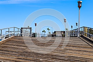 A long brown wooden pier with a gray metal hand rail and tall lamp posts along the pier surrounded by deep blue ocean water