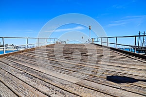 A long brown wooden pier with a gray metal hand rail and tall lamp posts along the pier surrounded by deep blue ocean water