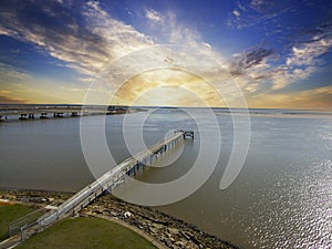 A long brown wooden pier of the rippling waters of Mobile Bay with cars driving on the highway and powerful clouds at sunset