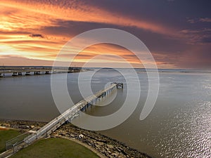 A long brown wooden pier of the rippling waters of Mobile Bay with cars driving on the highway and powerful clouds at sunset