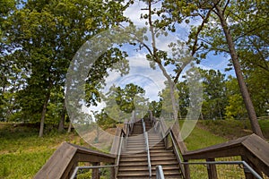 A long brown wooden staircase up a hillside surrounded by lush green trees, grass and plants with blue sky and clouds