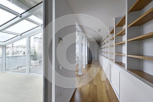 Long corridor of a penthouse apartment with a glazed terrace and a long empty shelf with wooden shelves and loose oak flooring
