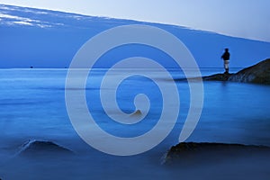 Long exposure of blue sea scape at morning light with man fishing on rock