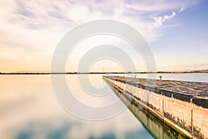 Long exposure of a bridge at evening before sunset. Blue sky, white clouds, sky mirrored in water, calm ocean