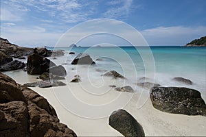 Long exposure of light waves rolling over large rocks in a idilic white sand beach in a small bay with small islands on