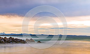 Long exposure shot of sandy beach shoreline against a backdrop of rocky outcrops and a cloudy sky.