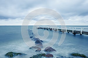 Long exposure shot of the sea with rocks and a pier on a cloudy day
