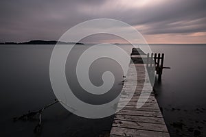 Long exposure view of a pier on a lake at dusk, beneath a dramatic, moody sky