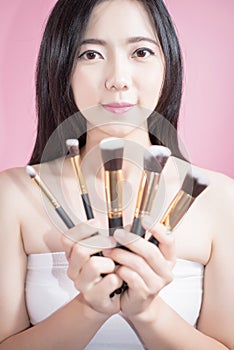 Long hair asian young beautiful woman smile and fun, touch her face and hold cosmetic powder brush set, isolated over pink.