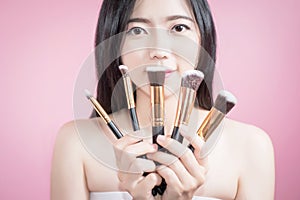 Long hair asian young beautiful woman smile and fun, touch her face and hold cosmetic powder brush set, isolated over pink.