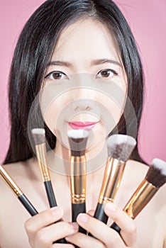 Long hair asian young beautiful woman smile and fun, touch her face and hold cosmetic powder brush set, isolated over pink.