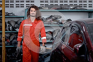 Long hair handsome mechanic man in red uniform standing around engine spare parts vehicle, auto mechanic technician inspecting