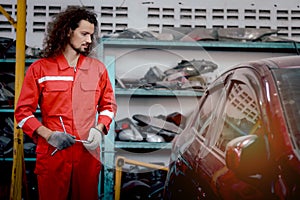 Long hair handsome mechanic man in red uniform standing around engine spare parts vehicle, auto mechanic technician inspecting
