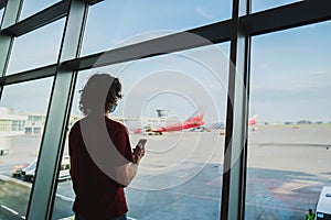 Long hair man in mask with smartphone in the airport with plane on the background