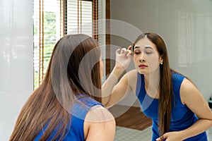 A long-haired Asian woman in a blue dress looks in the mirror with false eyelashes in her home bathroom