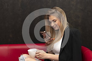 A long-haired blonde 20-25 years old with a mobile phone and a white cup on a black background.