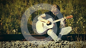 Long-haired blonde guitarist plays sitting on the railway on a sunny day