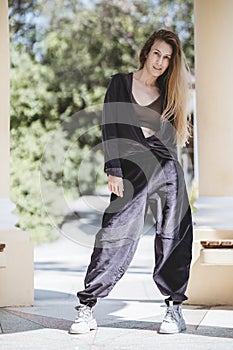 A long-haired blonde in wide pants and a shirt tied with a knot stands between the columns in the park on a sunny day