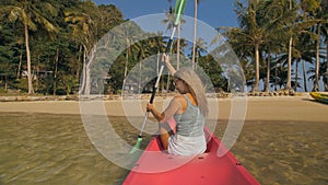 Long haired blonde woman with sunglasses rows bright pink canoe along sea bay water to beach with growing palms.