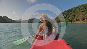 Long haired blonde woman with sunglasses rows bright pink canoe along sea bay water to beach with growing palms.