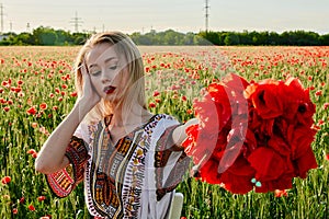 Long-haired blonde young woman in a white short dress on a field of green wheat and wild poppies