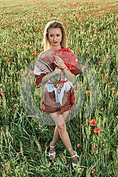 Long-haired blonde young woman in a white short dress on a field of green wheat and wild poppies