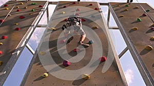 A long-haired boy skillfully climbs a rock-climbing wall in the open air, mastering heights and his own limits.