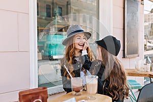 Long-haired brunette little girl gently touching mother`s face, sitting in outdoor cafe with glass of milk shake