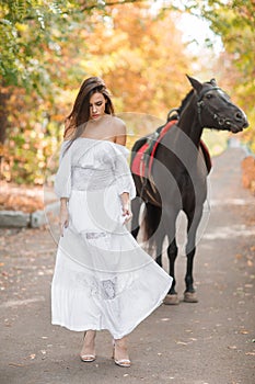 The long-haired brunette in a long dress is walking in front of a horse in the park.