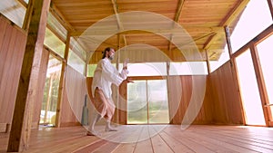 A long-haired Caucasian man in light and loose clothing practices qigong tai chi in a wooden practice room in summer