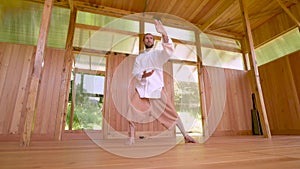 A long-haired Caucasian man in light and loose clothing practices qigong tai chi in a wooden practice room in summer