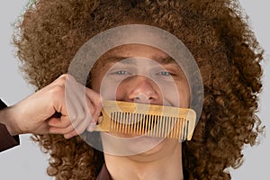 A long-haired curly-haired guy in a brown shirt on a gray background uses a wooden comb. Emotions before a haircut in a