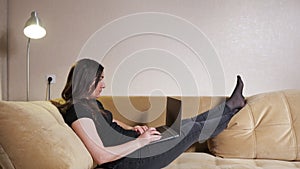 Long haired lady lies on brown sofa and types on grey laptop