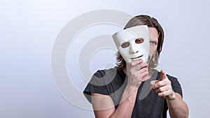 A long-haired man in a gray T-shirt looks out from behind a white mask and points his finger at the camera. The concept is you