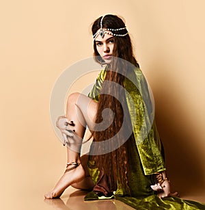 Long-haired model in green cape, jewelry on head, hands and leg. She sitting sideways on floor against beige background. Close up