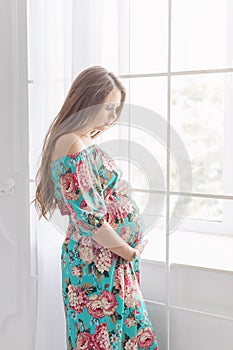 Long haired pregnant woman in white interior standing near the window.