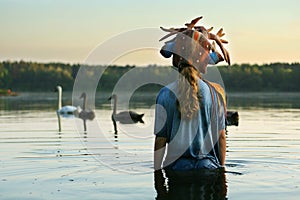 Long haired teenage girl wearing Antler bambi hat is staying waist-deep in the water watching swans family