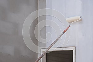 Long handle roller brush applying primer white paint with door frame on cement wall inside of house construction site, building